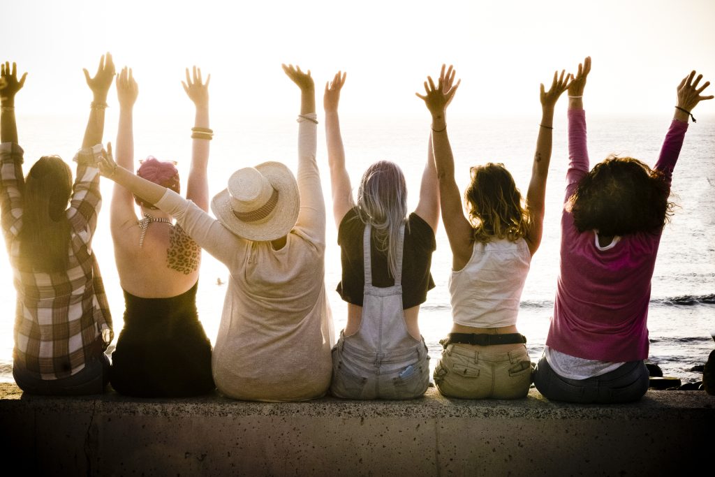 The backs of six women sitting on a beach wall celebrating with their hands in the air.